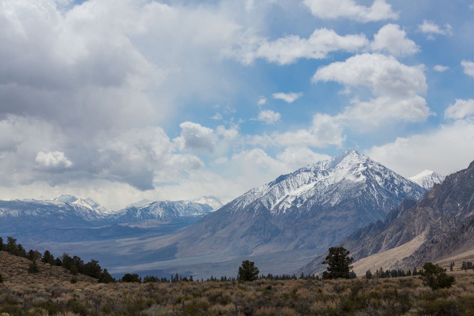A snow-capped Sierra Nevada peak under white clouds, with pines and sagebrush in the foreground