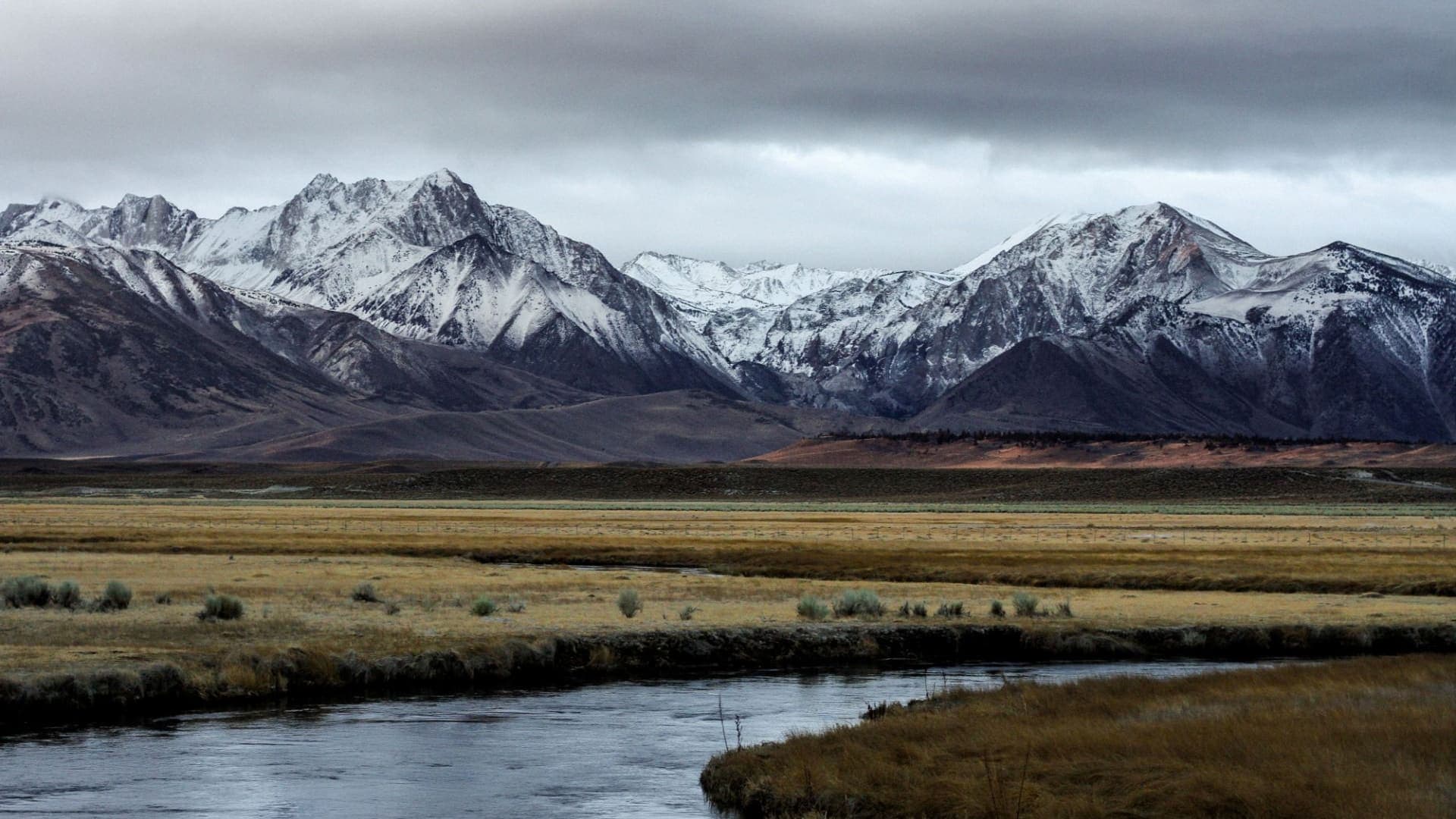 Snow-dusted Sierra Nevada peaks above a meadow and a winding stream under a gray sky