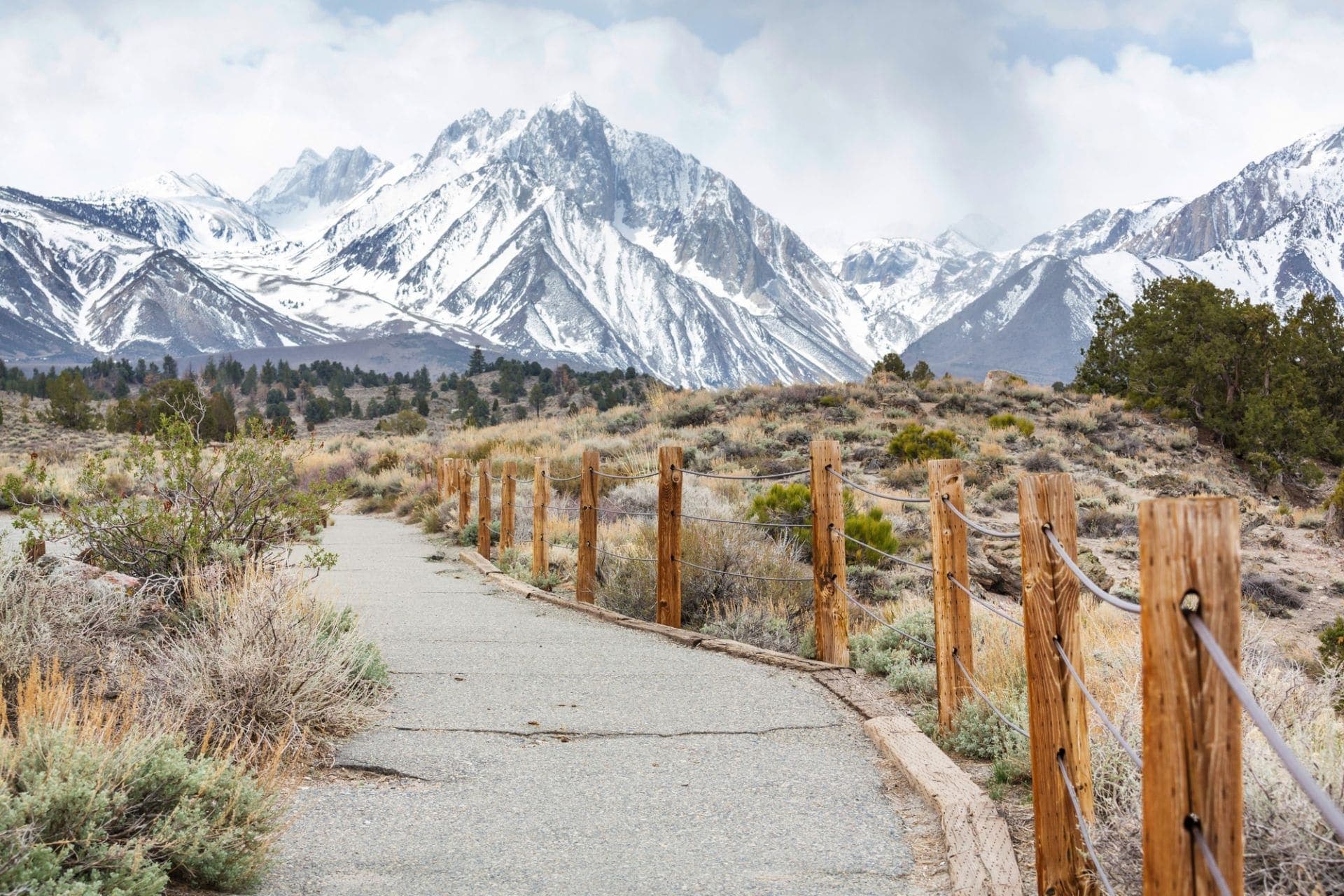 A paved trail beside a wooden post-and-rope fence, leading toward snow-covered Sierra Nevada peaks